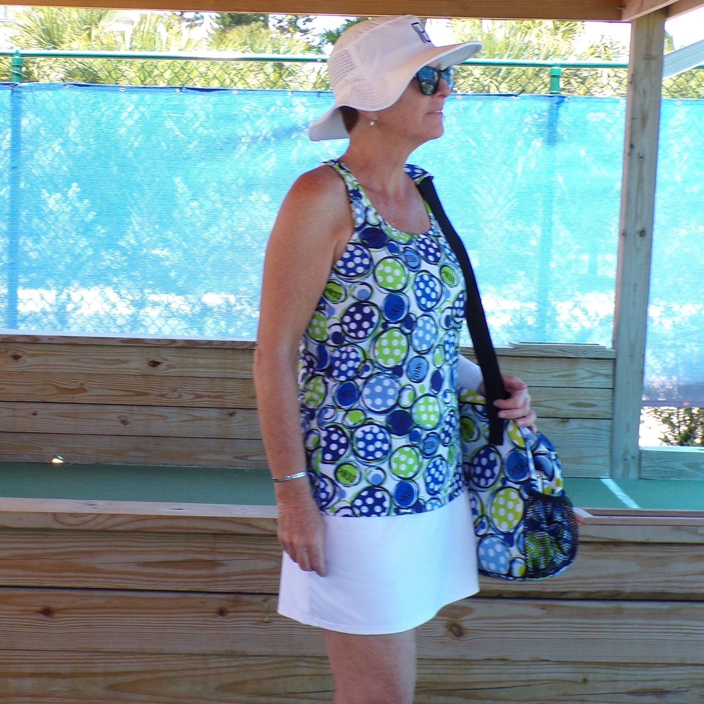 A woman stands sideways on a wooden deck in the Pickleball Bella PB Racerback Tank Dink 1, featuring a blue and green circular pattern, paired with a white skirt and bag. She wears a white sunhat and sunglasses before blue mesh fencing.