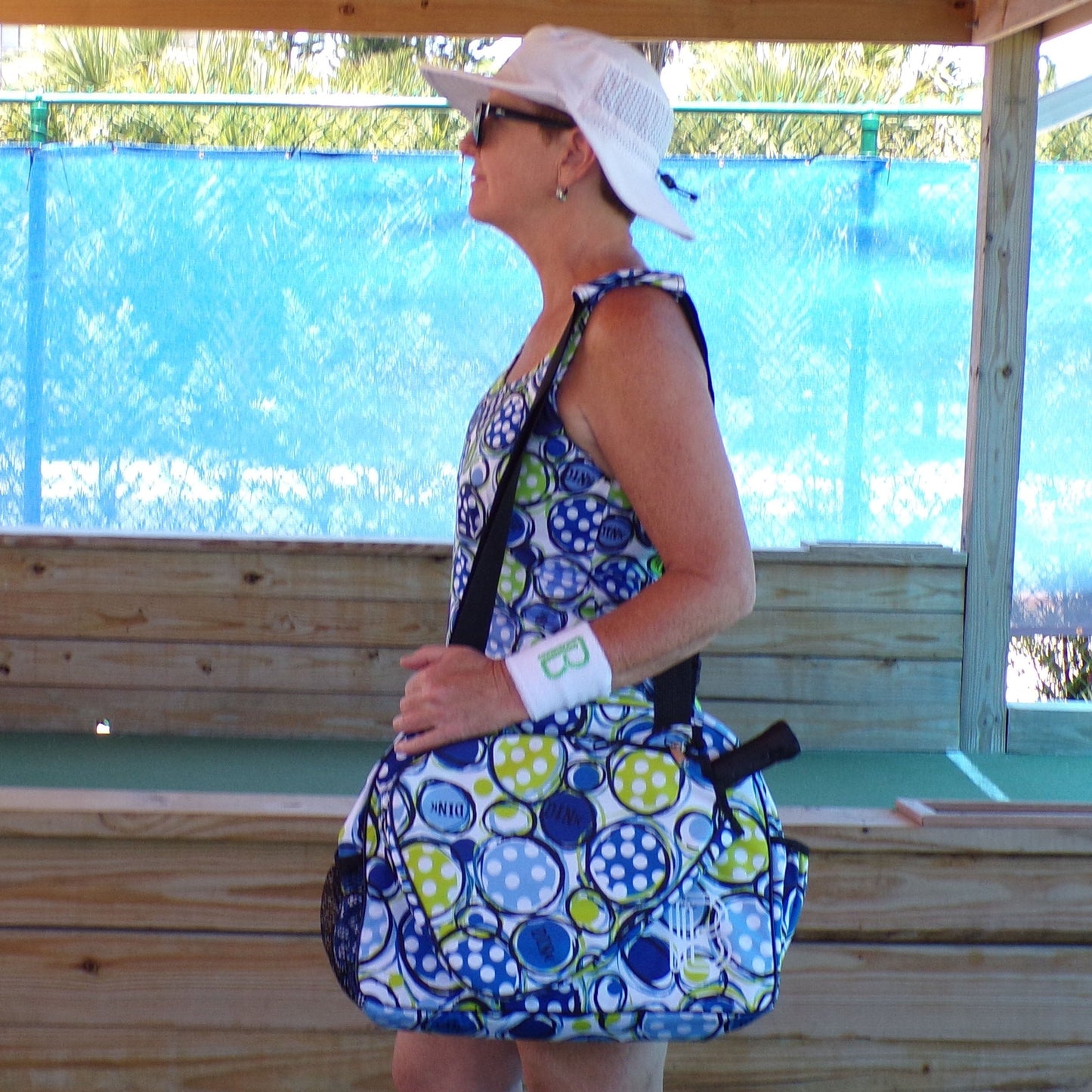 A woman in profile wears the Pickleball Bella PB Racerback Tank Dink 1, a patterned performance dress, with a white visor, sunglasses, and "B" wristband. She carries a large matching bag, standing near wood and blue mesh outdoors.