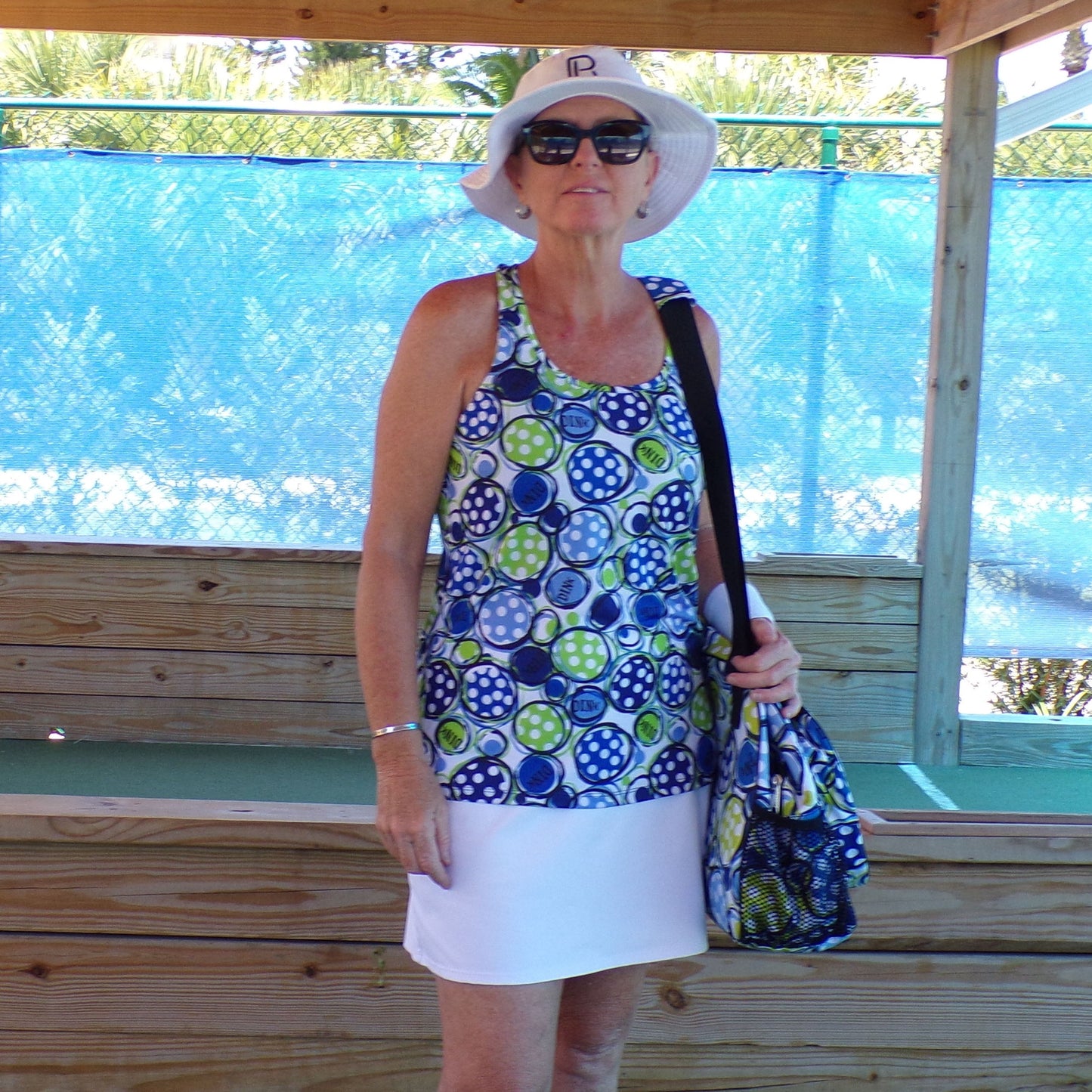 A woman stands outside by a wooden bench and blue mesh fence, wearing dark sunglasses, a white sun hat, and the Pickleball Bella PB Racerback Tank Dink 1 with green and blue circles, paired with a white skirt and matching bag.