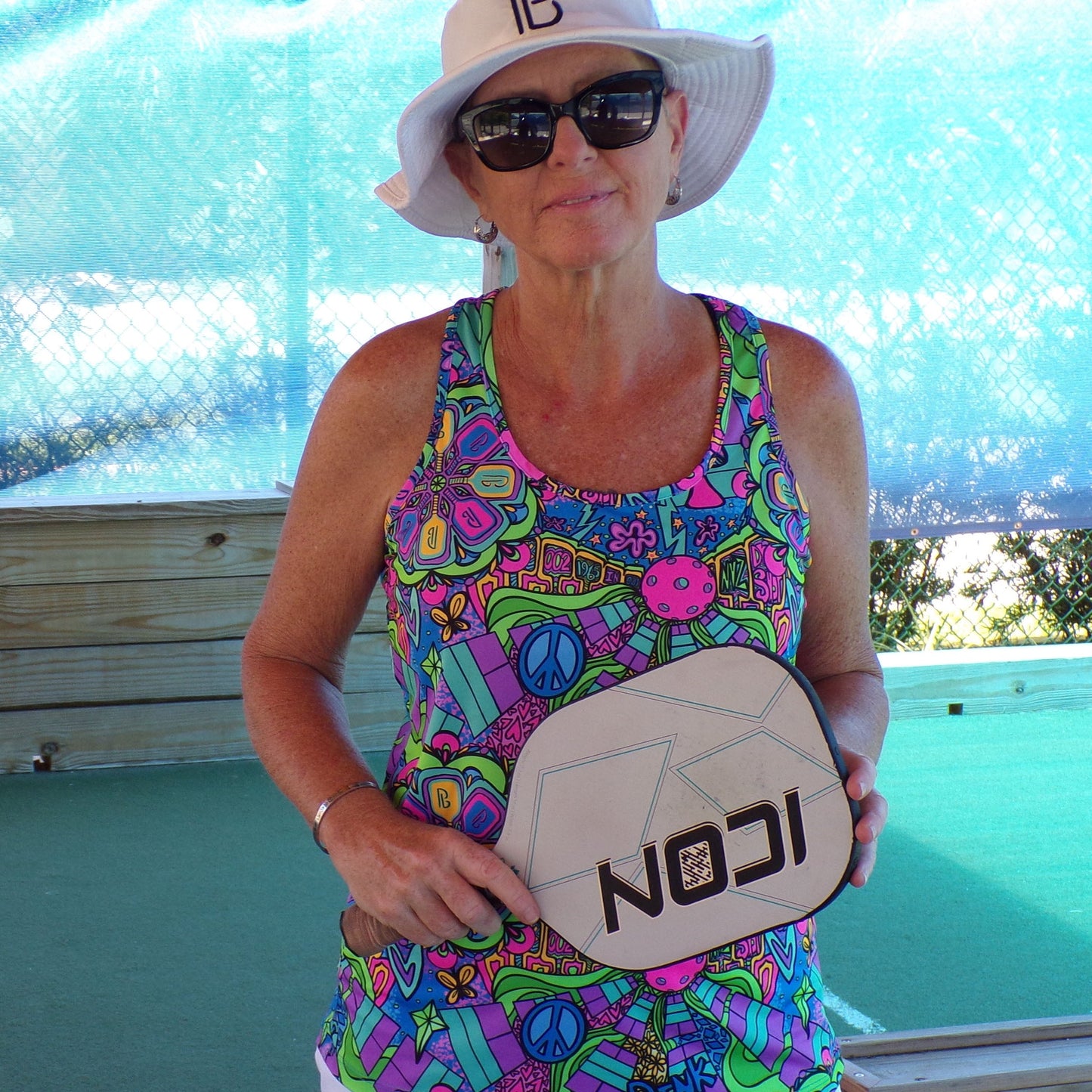 A woman in the Pickleball Bella PB Racerback Tank Groovy and sunglasses holds a beige ICON paddle by a wooden barrier on a green court, ready for action in her colorful performance top.
