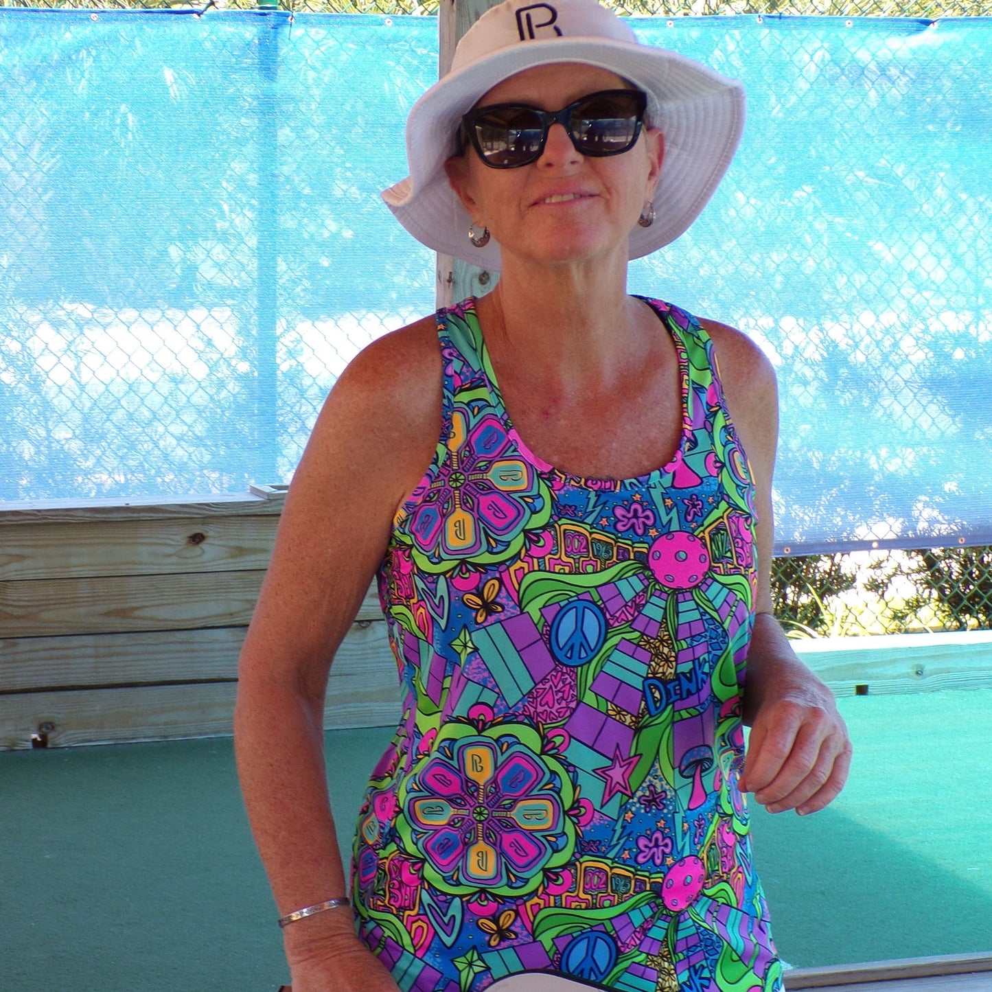 A woman in a Pickleball Bella PB Racerback Tank Groovy stands outdoors by a wooden fence, wearing a white sun hat and black sunglasses. Blue mesh and chain-link fences are visible in the background on this sunny day.