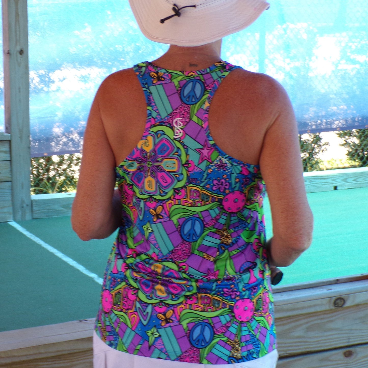 A person stands with their back to the camera wearing the Pickleball Bella PB Racerback Tank Groovy, featuring peace signs, flowers, and vibrant patterns. They pair it with a white sun hat and shorts as sunlight filters through greenery behind them.