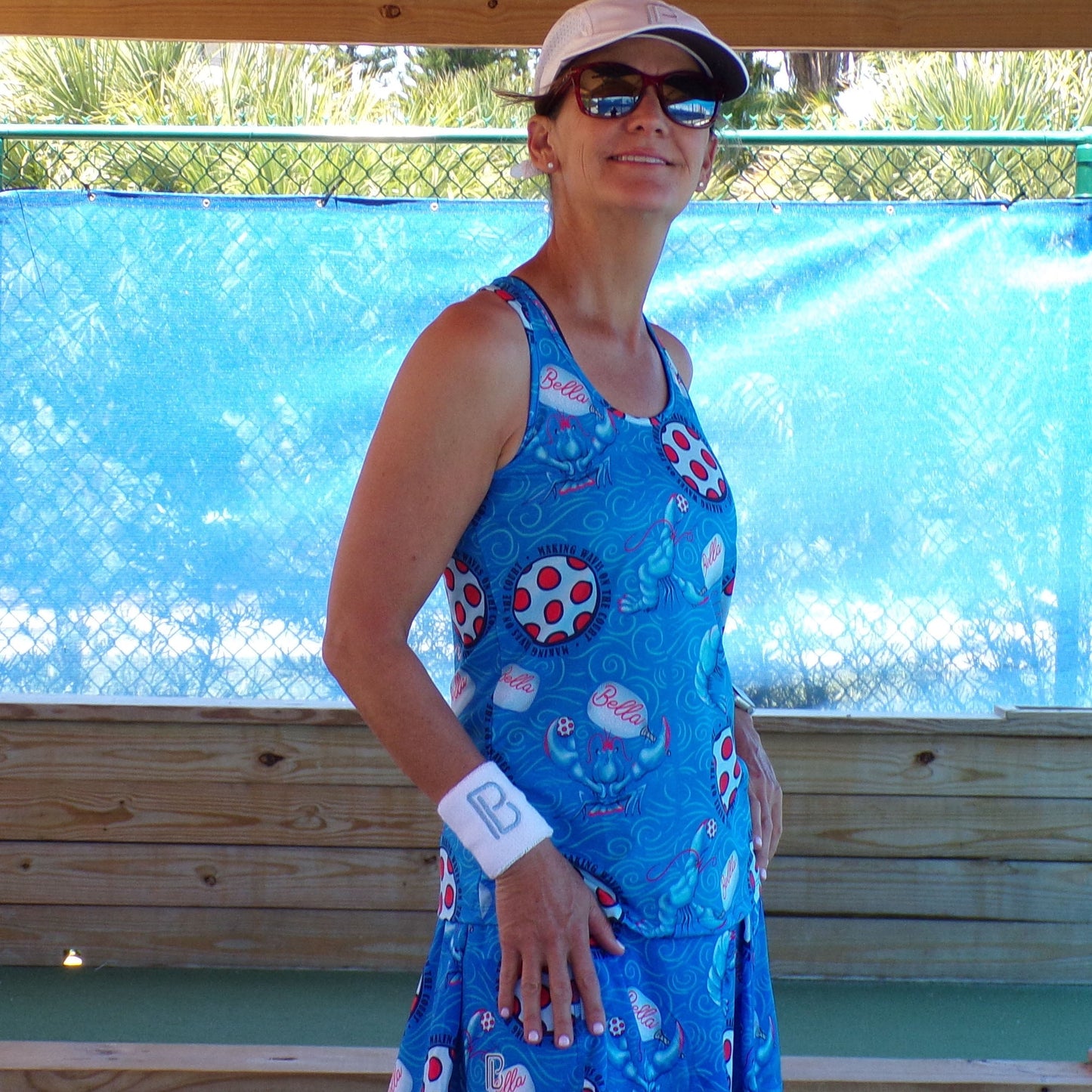 A woman smiles on a wooden deck, wearing Pickleball Bella's PB Racerback Tank Lobster Monster, a white visor, sunglasses, and a "B" wristband. A blue mesh fence and green trees are in the background.