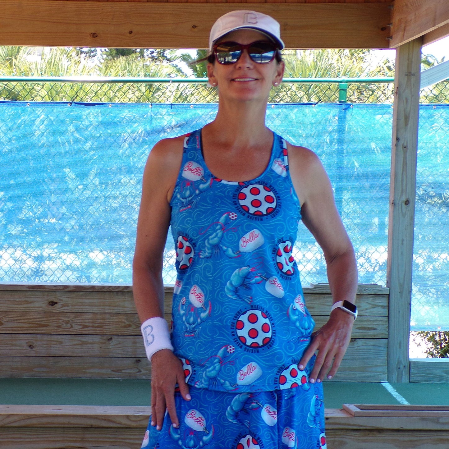 A woman stands outdoors smiling with her hand on her hip, wearing the Pickleball Bella PB Racerback Tank Lobster Monster, a white visor, sunglasses, and a “B” wristband in front of a blue mesh fence and wooden structure.