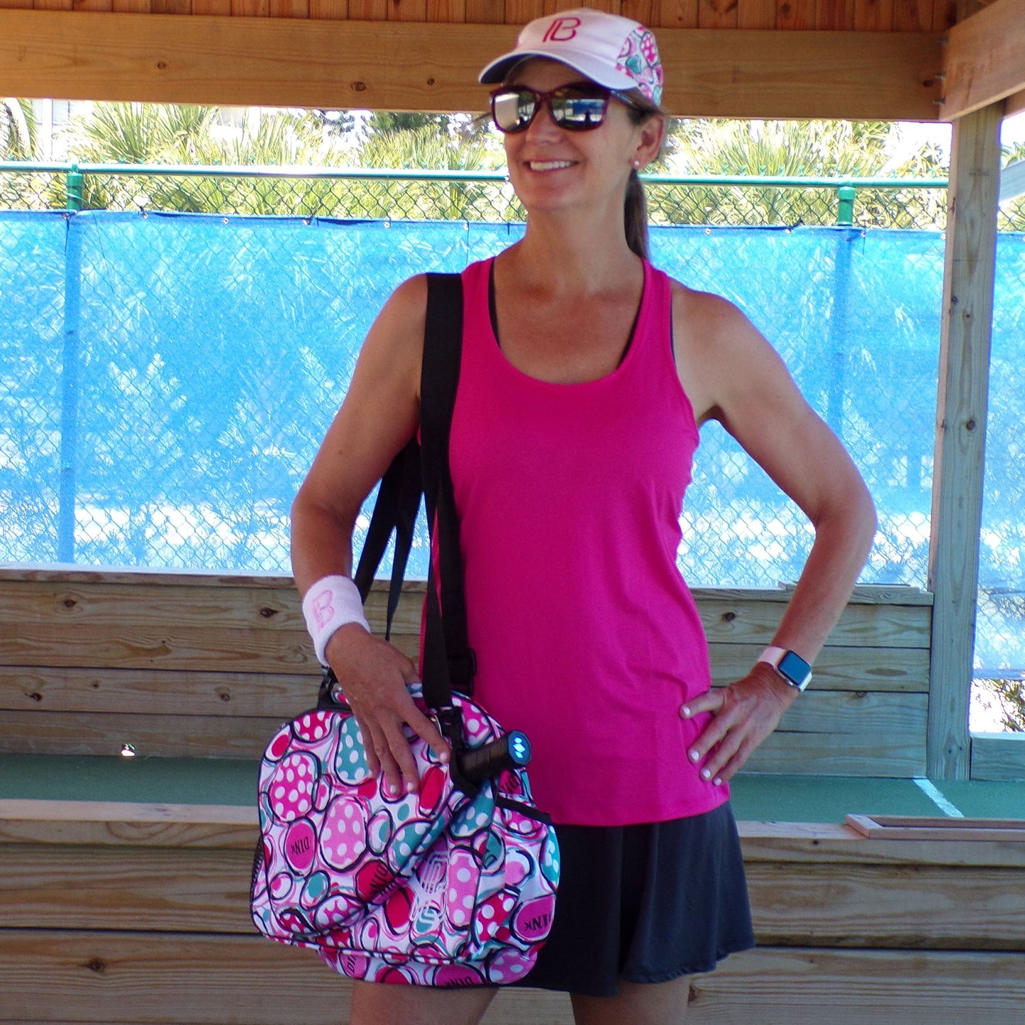 A woman smiles under a wooden shelter in a Pickleball Bella PB Racerback Tank Pink with SPF 50+ UV protection, black skirt, white wristband, sunglasses, pink-patterned cap, matching bag with blue water bottle, and a smartwatch.