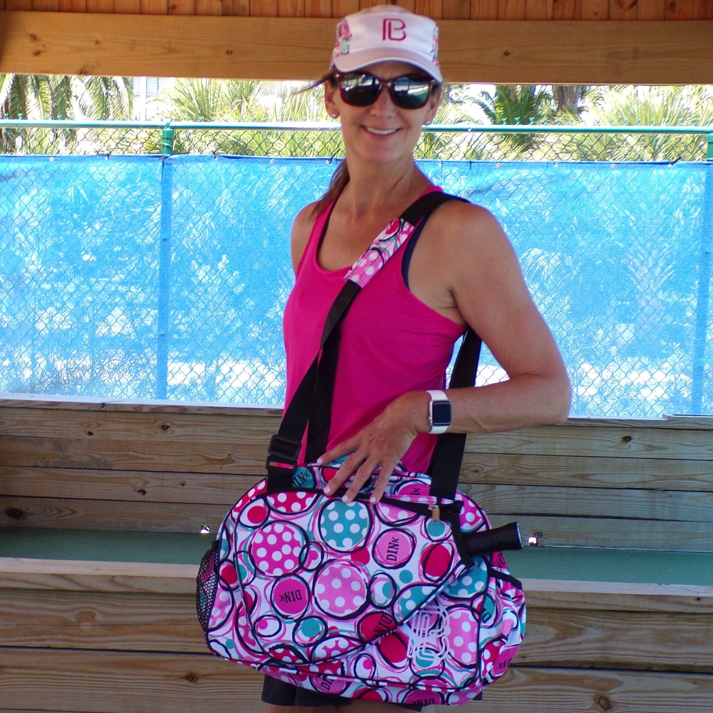 Wearing the Pickleball Bella PB Racerback Tank in pink (SPF 50+ UV protection), a woman smiles with a white hat and sunglasses, holding a polka-dot sports bag before a wooden bench, shaded roof, palm trees, and blue fence.