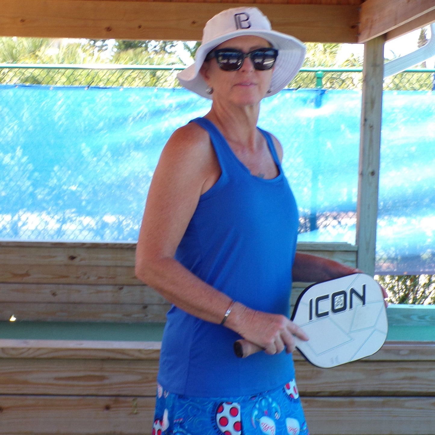 A woman wearing the Pickleball Bella PB Racerback Tank in Royal Blue with SPF 50+ stands on a pickleball court, holding an ICON paddle and facing the camera. She pairs it with a patterned skirt, white sun hat, and sunglasses.