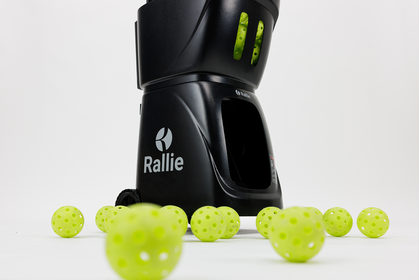 The Rallie Pickleball Machine - Midnight Black by Rallie Pickleball sits upright on a white surface, wheels visible, surrounded by bright yellow-green perforated balls—a portable, efficient solution for pickleball training anywhere.