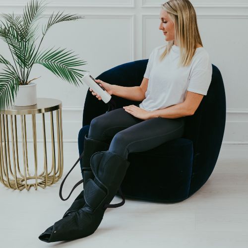 A woman in a white t-shirt and black leggings sits in a navy blue chair using the iReliev Relaxable Leg & Foot Air Compression System, holding its remote. A plant is on a gold side table nearby against a white wall.