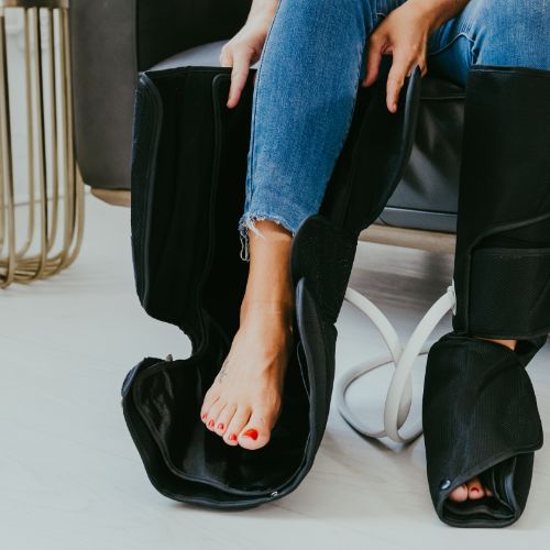 A person sits on a chair, wearing blue jeans and bare feet, using the iReliev Relaxable Leg & Foot Air Compression System. The open black sleeves connect with white tubes, and one sleeve reveals their red-painted toenails.