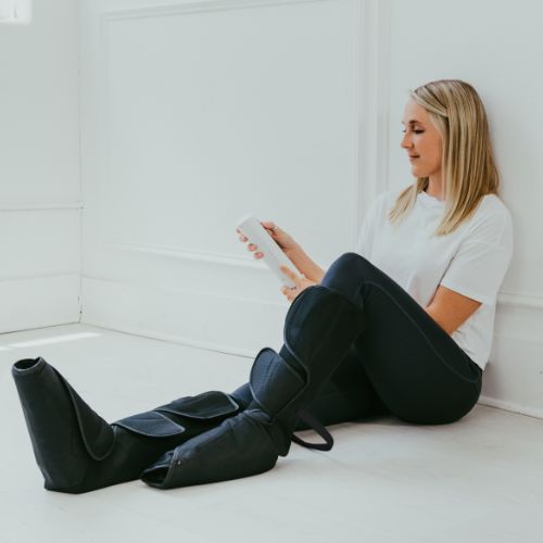 A woman with blonde hair sits on the floor in a white shirt and black pants, using the iReliev Relaxable Leg & Foot Air Compression System. She holds the white remote and looks at it with a relaxed expression.