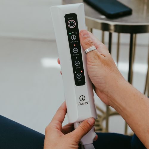 A person holds an iReliev Relaxable Leg & Foot Air Compression System, featuring a white body, black control panel, buttons, and indicator lights. They wear a silver ring and sit by a metallic table with a phone in the background.