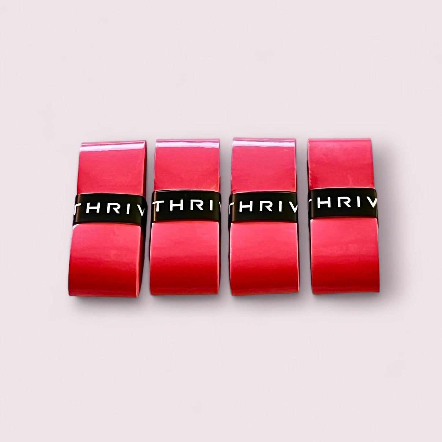 Four red rectangular lipstick tubes stand upright on a pale pink background, each with a black center band resembling Thrive Pickleball Overgrips (Super Tacky) and “THRIV” partially visible across the tubes.