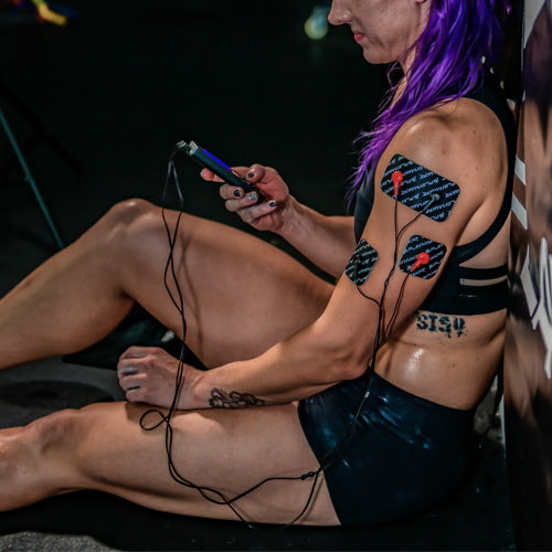 A woman with purple hair in athletic wear sits on the floor, sweaty from exercise, holding the iReliev SPORT Muscle Stimulator with Explosive Strength Mode. Wires connect to electrodes on her upper arm, and a side tattoo is visible.
