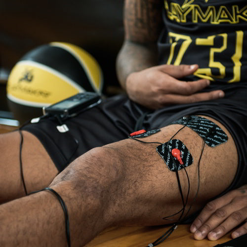 Wearing black athletic shorts and a basketball jersey, a person uses the iReliev SPORT Muscle Stimulator with Explosive Strength Mode with electrode pads on their thigh and knee, while a basketball sits nearby on the wooden floor.