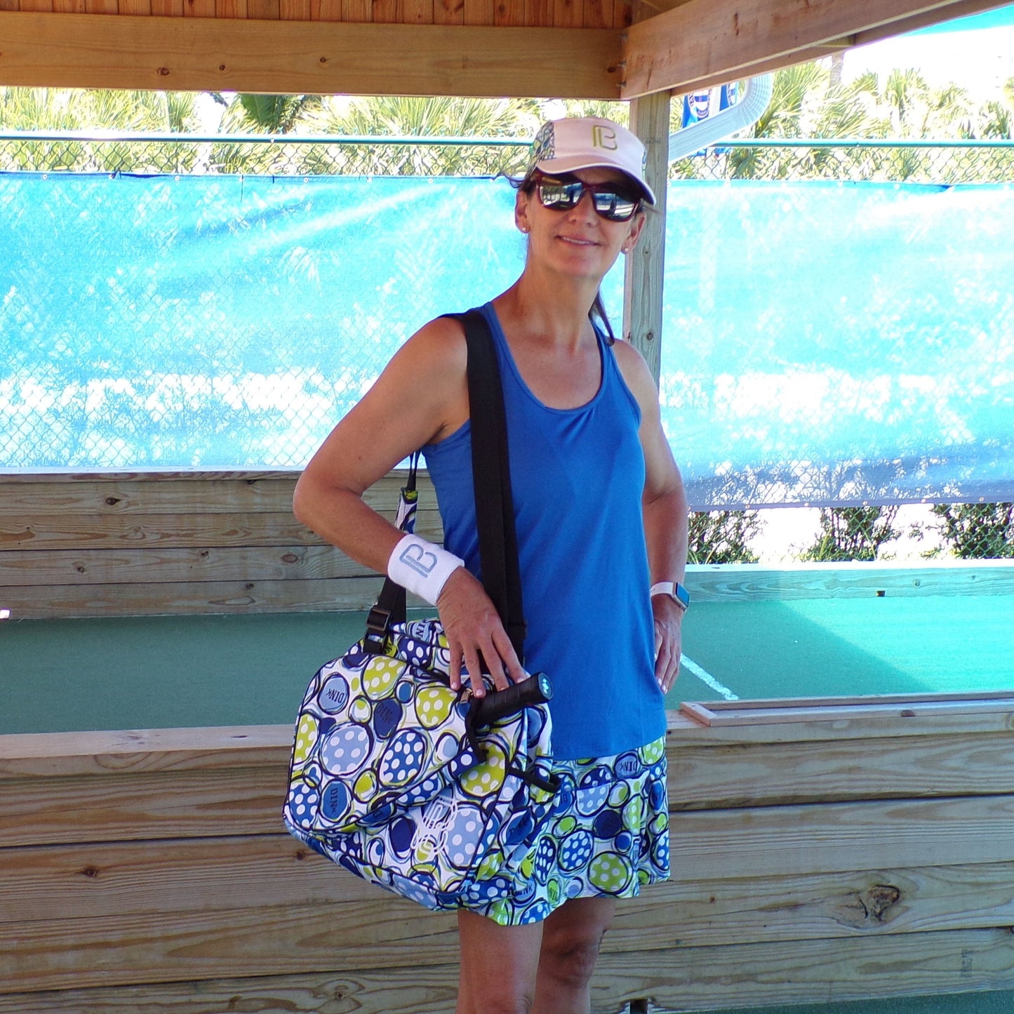 A woman wears the Pickleball Bella PB Racerback Tank in Royal Blue with SPF 50+ on a shaded pickleball court, paired with a patterned skirt, white visor, sunglasses, wristbands, and colorful bag. Palm trees are visible in the background.