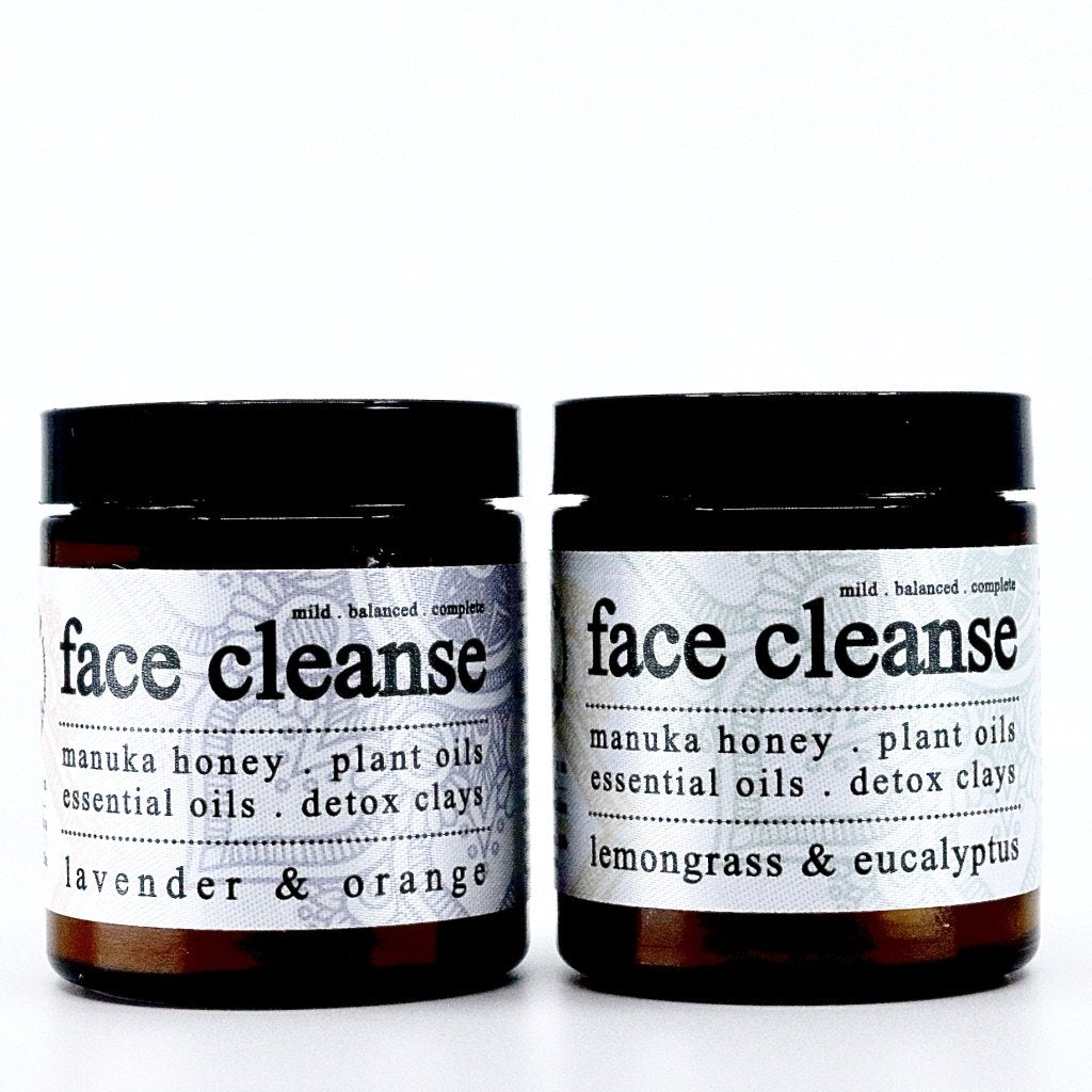 Two amber glass jars of Roots Apothecary face cleanse sit side by side on a white background. The left jar is lavender & orange, the right is lemongrass & eucalyptus. Both feature manuka honey, plant oils, and detox clays for a refreshing cleanse.