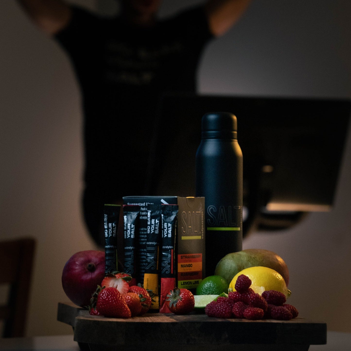 A dark, moody scene features assorted fruits on a wooden tray. Nearby are SALT Electrolyte Drink Mix Single-Serve Mixes for Hydration and Recovery by SALT Electrolytes and a tall black SALT bottle, with a blurred figure in the background.