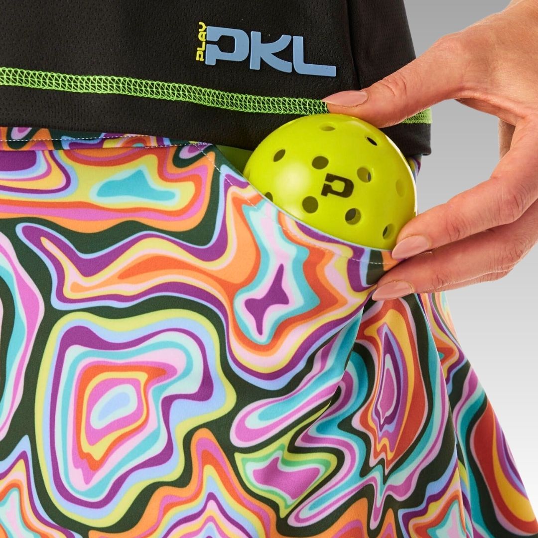 A person puts a yellow pickleball with black dots into the pocket of the colorful, swirling-patterned Sammy Skort from Playly Store. The PLAY PKL logo is on the waistband, and its sofKnit fabric provides comfort against a gray background.
