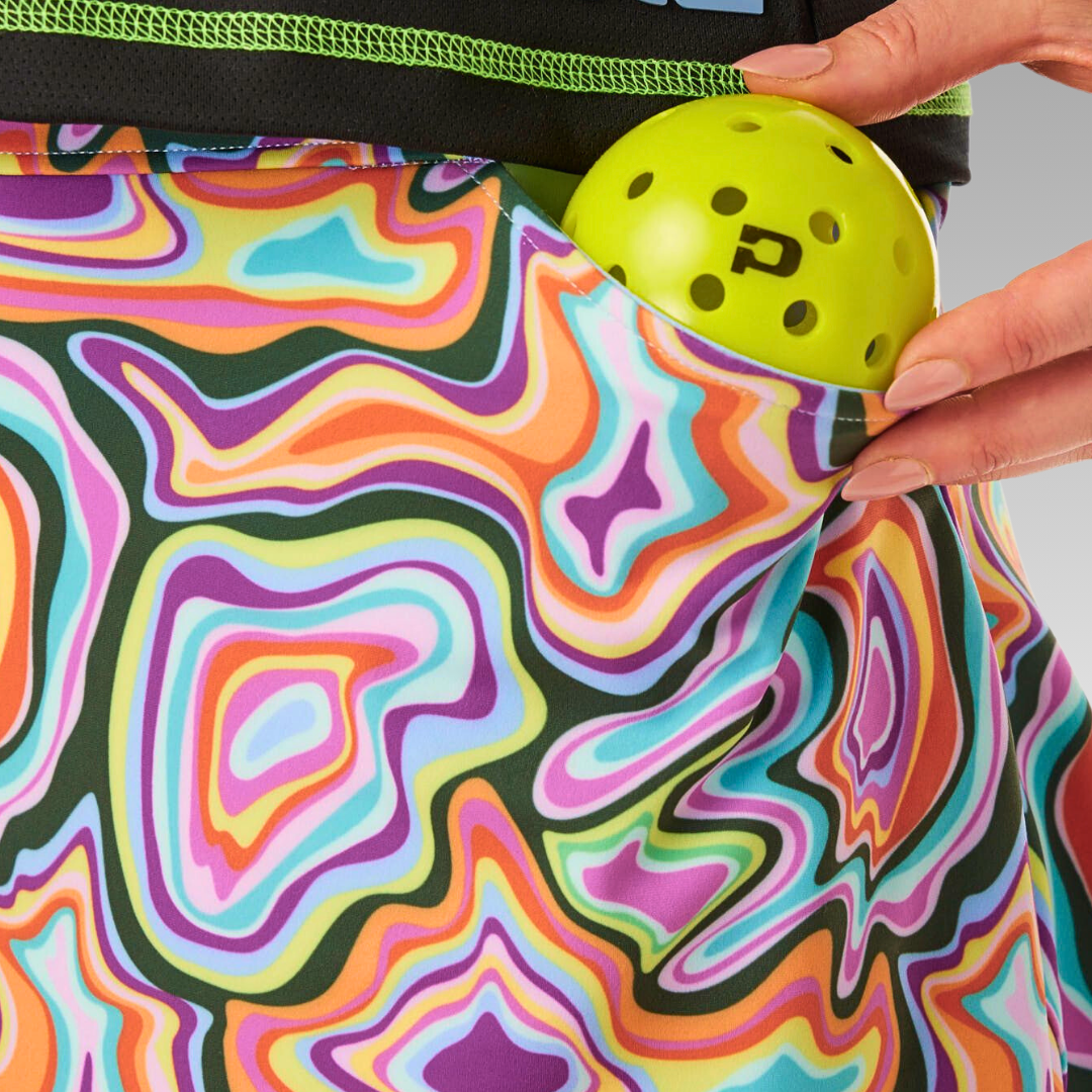 A hand slips a yellow P-logo pickleball into the pocket of Playly Store's Sammy Skort 2.0, featuring a colorful wavy pattern and crafted from CoolFlow fabric, against a plain gray background focusing on the lower torso and hand.