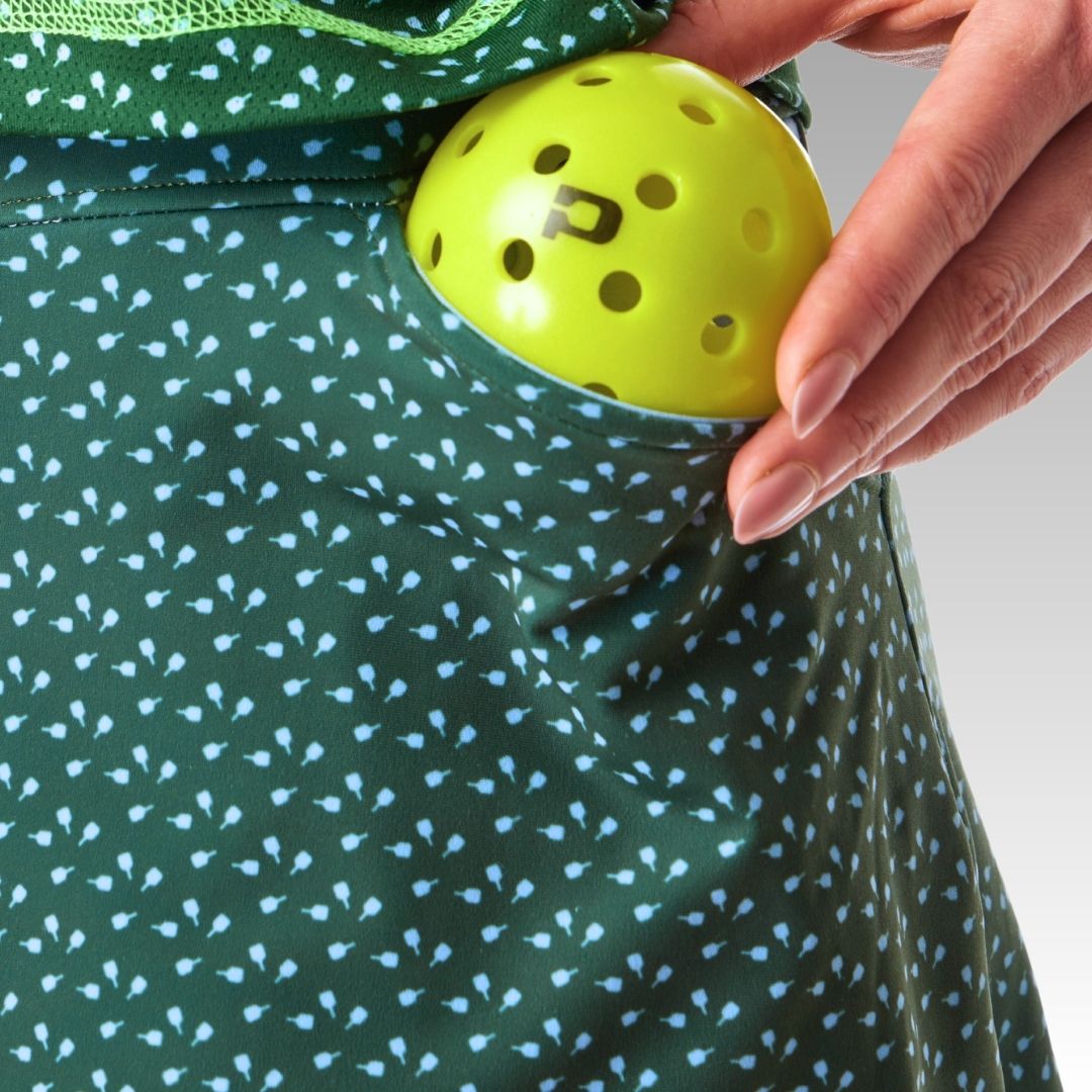 A close-up of a hand placing a yellow ball into the side pocket of the green Sammy Skort by Playly Store, featuring light blue floral prints and moisture-wicking SofKnit fabric. The background is a neutral gray gradient.