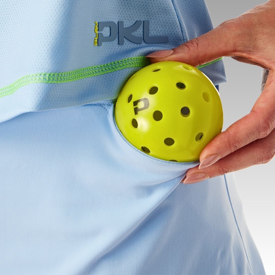 A hand puts a yellow pickleball with black holes into the side pocket of the light blue Playly Store Sammy Skort, made from SofKnit fabric with green trim, moisture wicking, and "PKL" above the pocket on a gray gradient background.