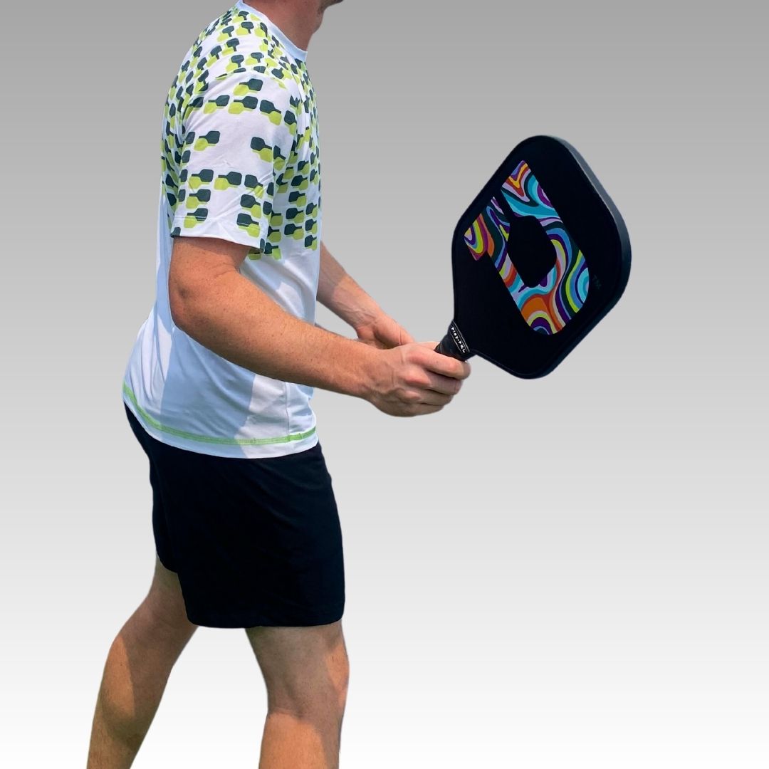 A person in a white and green patterned shirt and black shorts holds the Playly Store Signature Pickleball Paddle, featuring a colorful graphite surface. The image is cropped from shoulders up and below the knee, set against a smooth gray gradient background.