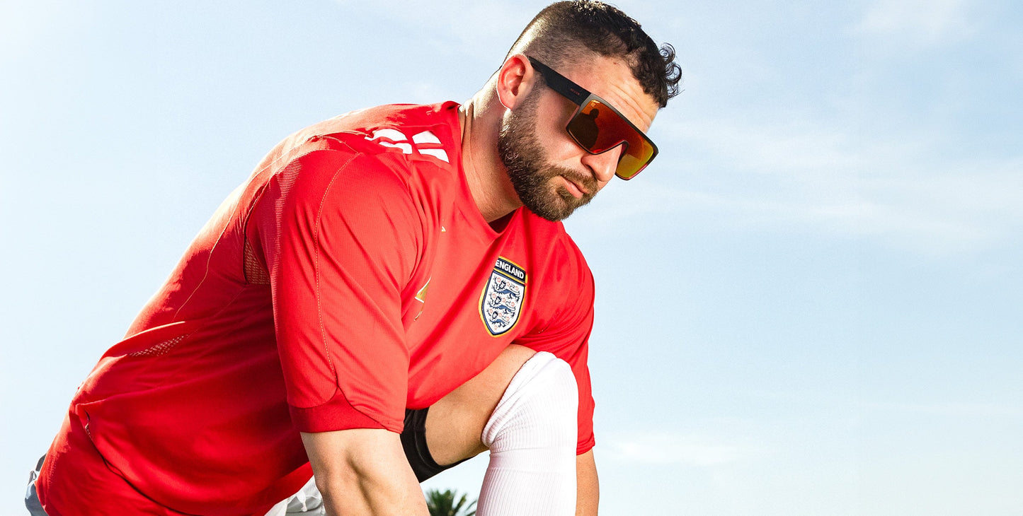 A bearded man wearing Neven Eyewear's 2nd Light sunglasses with UV400 protection kneels outdoors under a blue sky, focused as he ties his shoe. He sports a red England jersey with the national team logo and white stripes.
