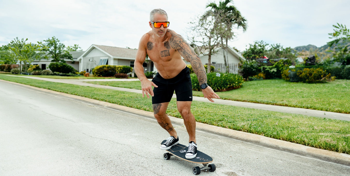 A shirtless, tattooed gray-haired man rides a skateboard down a suburban street, wearing Neven Eyewear's 2nd Light red polarized sunglasses, black shorts and sneakers, with green grass and trees bordering single-story houses under cloudy skies.
