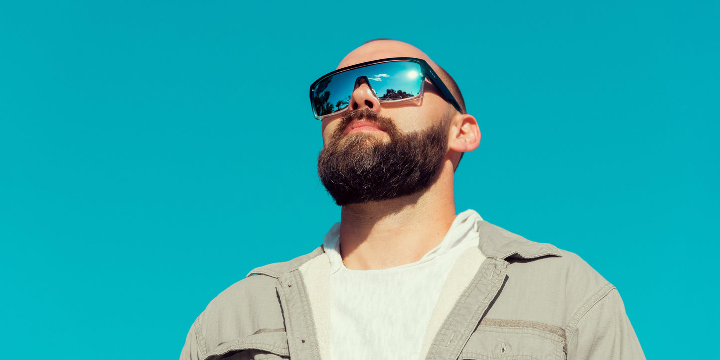 A bearded man in a white t-shirt and light gray button-up stands confidently against a blue sky, wearing Neven Eyewear’s Inlet sunglasses with impact-resistant, polarized UV400 lenses that reflect trees as he gazes upward with a calm expression.