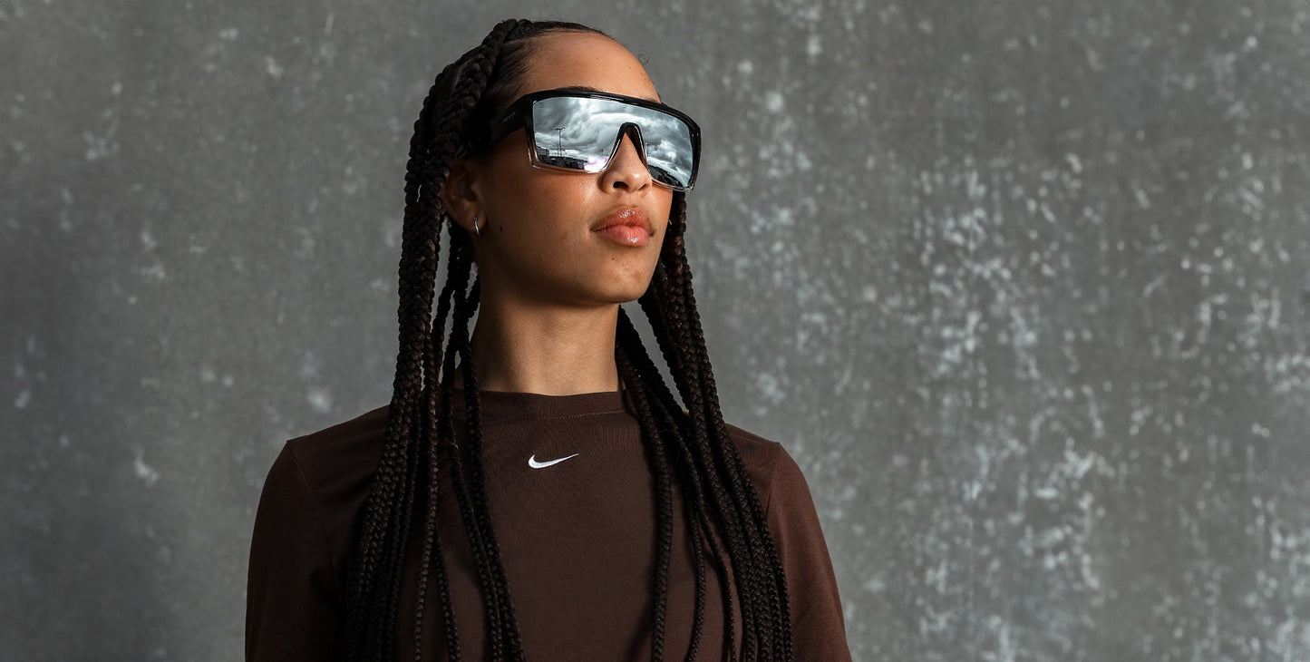 A woman with long braids wears Inlet polarized UV400 sunglasses from Neven Eyewear, paired with a brown Nike top. She stands against a gray, speckled background, confidently gazing sideways as her lenses reflect a cloudy Sebastian Inlet sky.