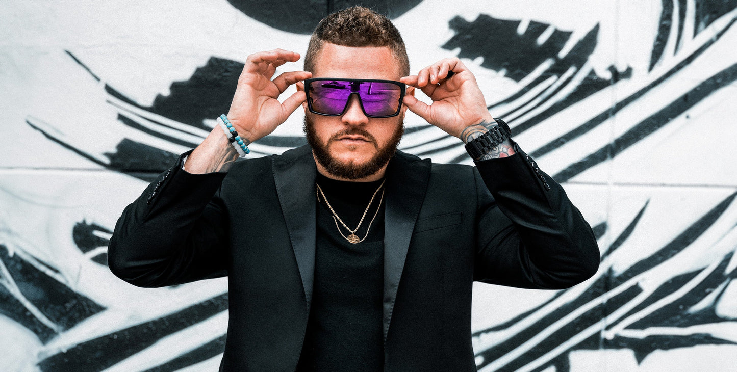 A bearded man in a black blazer and gold chains adjusts his Neven Eyewear Paradise sunglasses, which are large and reflective. Behind him is a bold black and white abstract mural with striking stripes and shapes.
