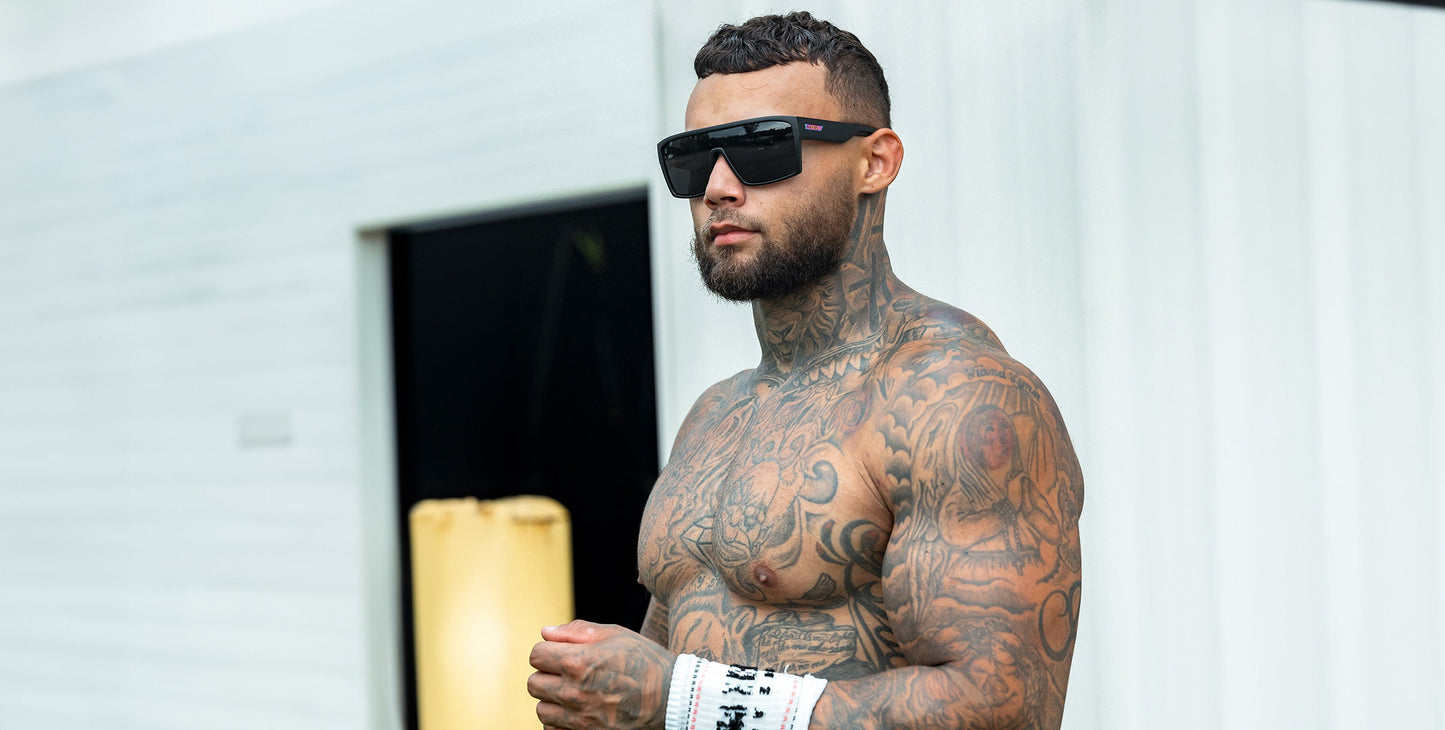 A muscular, tattooed man stands shirtless outdoors, wearing Neven Eyewear's RC's black polarized sunglasses with UV400 protection and white hand wraps. He has short dark hair, a trimmed beard, and a serious look. A white building is behind him.