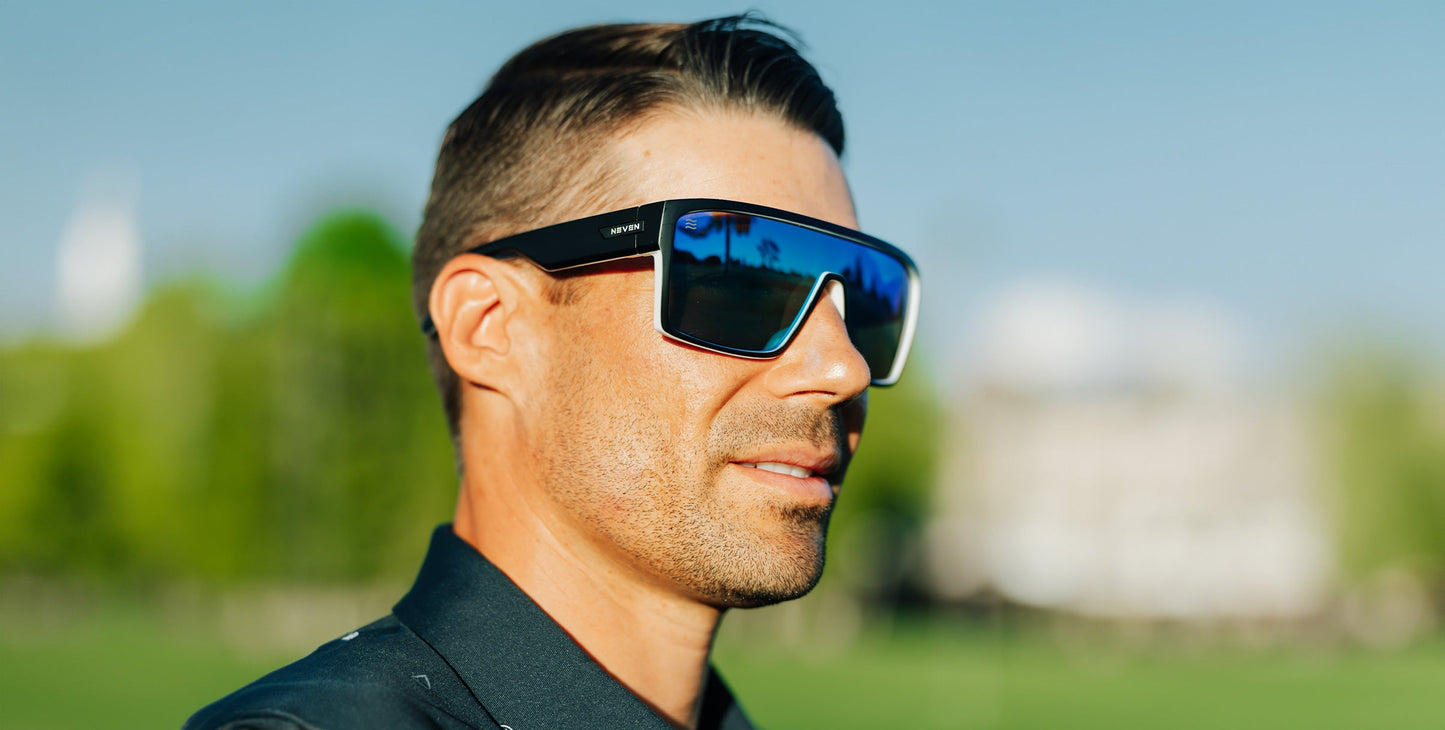 A man with short brown hair and stubble wears Neven Eyewear’s Spanish House oversized, polarized UV400 sunglasses and a dark collared shirt. He stands outdoors in bright sunlight, with grass and trees blurred in the background.