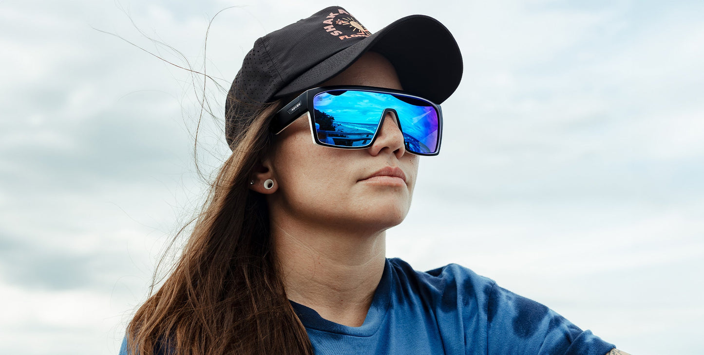A person wearing Neven Eyewear’s Spanish House polarized UV400 sunglasses and a black cap gazes into the scenic, cloudy sky as waves at Space Coast blow their long brown hair. Reflections dance on their shades in the breezy landscape.