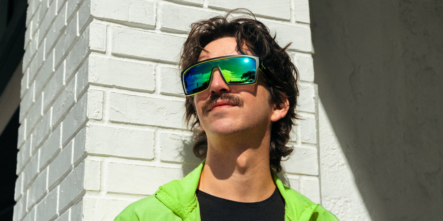 A man with wavy brown hair and a mustache wears Neven Eyewear's Tables sunglasses, featuring large polarized UV400 lenses, along with a bright green jacket as he stands in sunlight against a white brick wall, looking slightly upward.