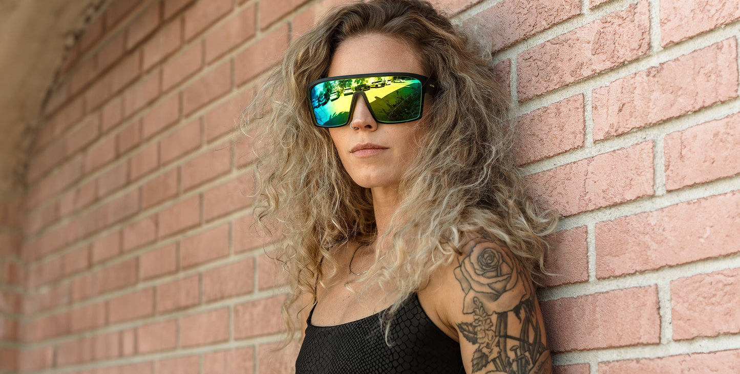 A confident woman with long curly blonde hair and a tattooed arm leans against a pink brick wall, wearing black sleeveless top and Neven Eyewear Tables sunglasses—large, mirrored, polarized lenses with UV400 protection.