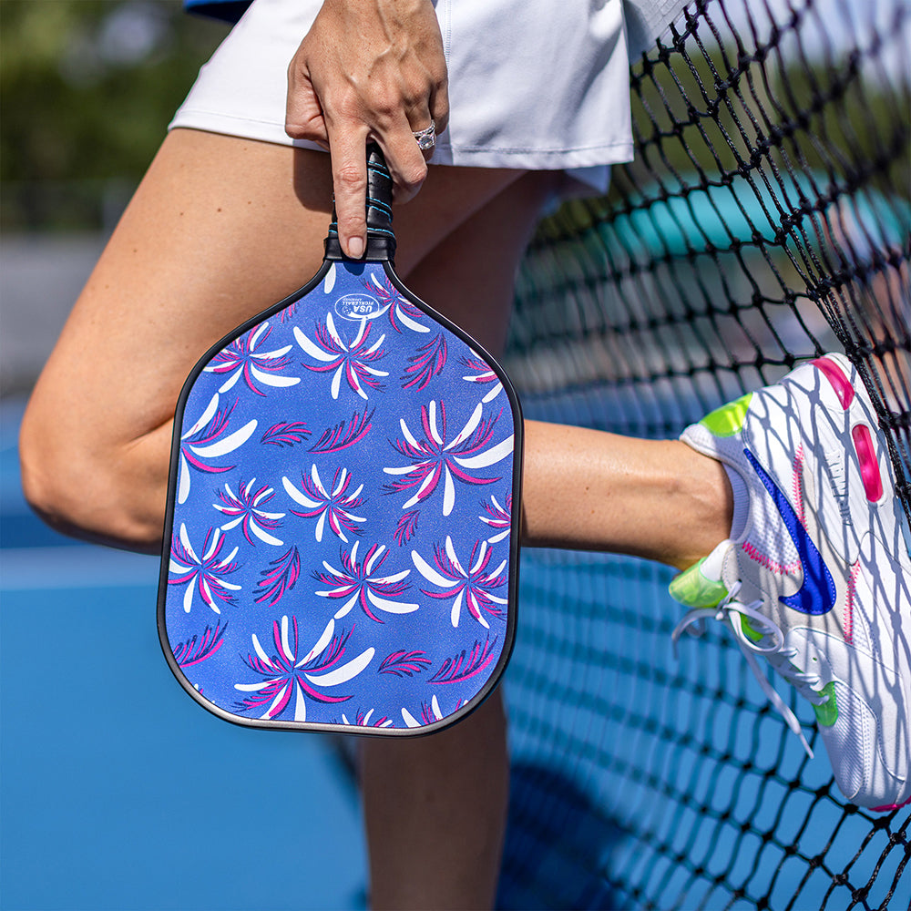 A person in a white skirt leans against a tennis net, holding the Swinton Pickleball Eclipse Island Vibes Pickleball Paddle featuring pink and white abstract flowers. Their shoes are white Nikes with colorful branding; one foot rests on the net.