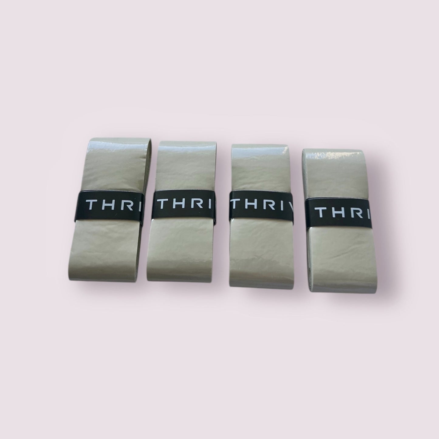 Four beige rectangular Thrive Pickleball Overgrips (Super Tacky) are lined up side by side on a light pink background, each folded and secured with a black band marked “THRI.” Durable and super tacky for reliable performance.