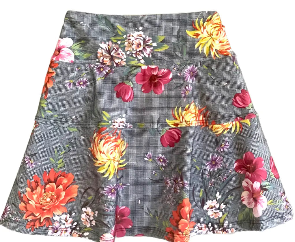 The Nola Plaid Floral Skirt by Runway Athletics is a knee-length A-line tennis skirt with a gray plaid base, colorful floral prints, three seams, a fitted waistband, and a moisture-wicking under-short for comfort.
