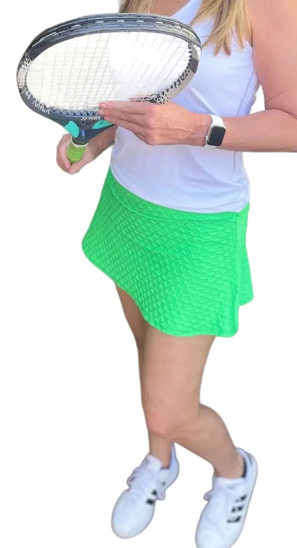 A woman wears a white sleeveless top, the Runway Athletics Nola Lime Scallop Skirt, white sneakers, and a smartwatch while holding a tennis racket. Only her body and part of her face are shown against a white background.