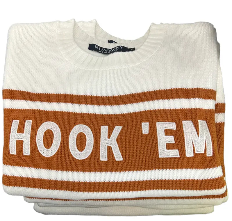 A folded white HOOK 'EM Tennis Sweater - Limited Edition by Runway Athletics features bold burnt orange stripes and "HOOK EM" across the front center, stacked neatly against a plain white background—a true display of Longhorn pride.