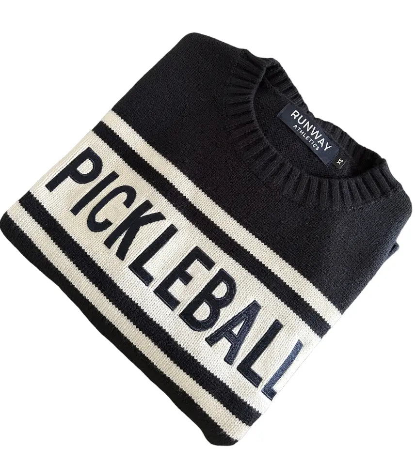 The Pickleball Sweater - Navy/Cream by Runway Athletics features a crew neck, two cream stripes, “PICKLEBALL” across the chest, and an acrylic weave. Folded neatly on a white background, it showcases the "RUNWAY" tag inside the collar.