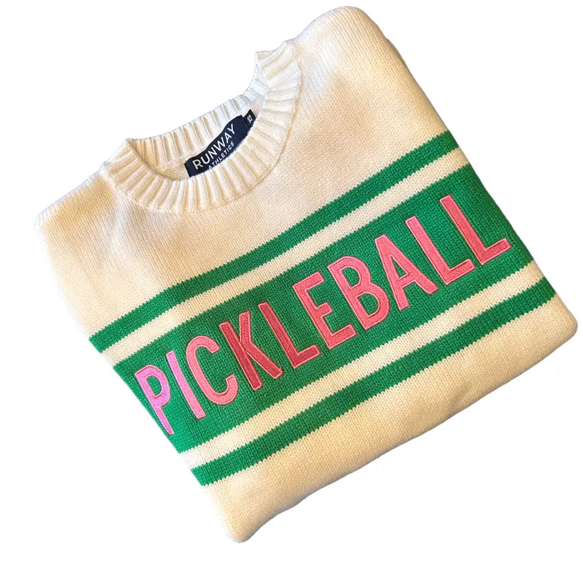 A folded cream crew neck Pickleball Sweater by Runway Athletics features green stripes and bold pink “PICKLEBALL” letters across the front, with the brand label visible by the collar, displayed on a white background.
