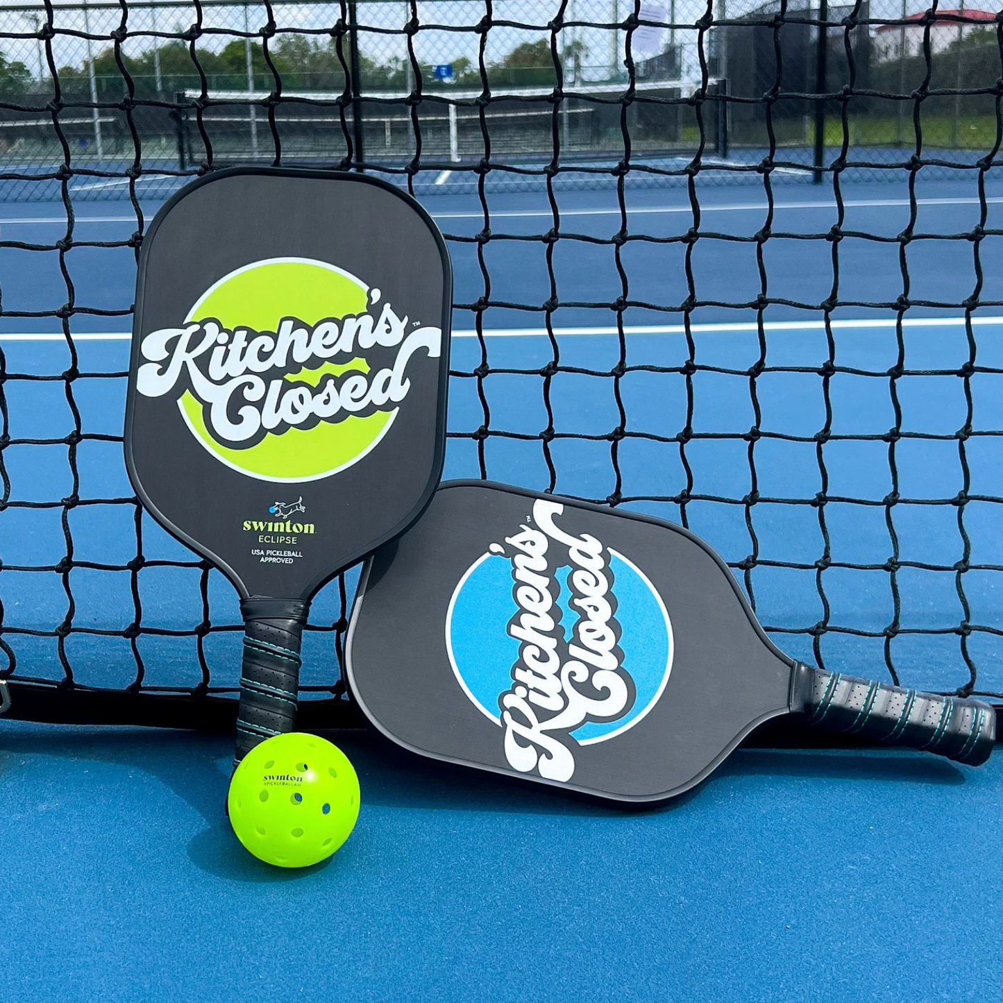 Two Swinton Pickleball Eclipse Kitchen's Closed paddles with textured graphite faces and black handles, plus a yellow ball, rest on a blue court with visible lines and fencing against a black net.
