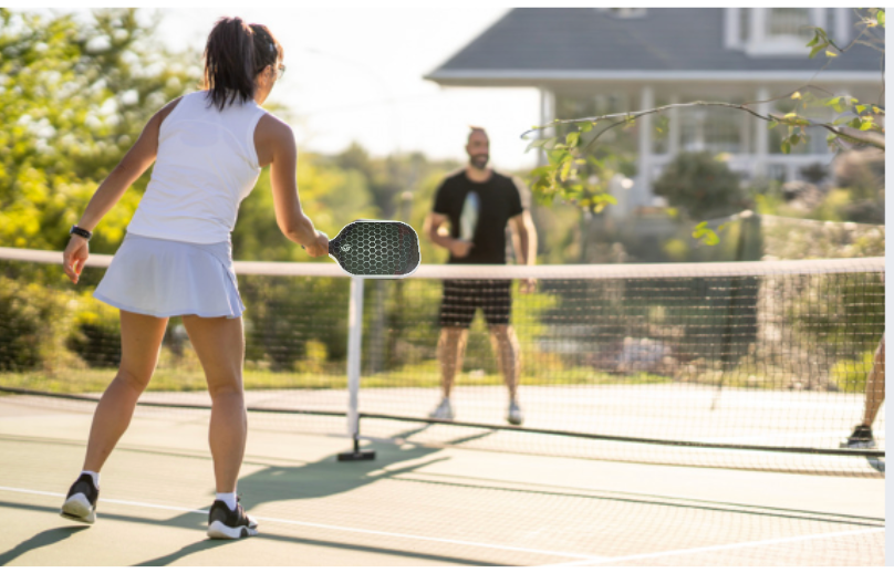 A woman in white sportswear gets ready to hit a ball on an outdoor court, using The Big Dink - Portable Pickleball Net by The Big Dink, while a man prepares on the opposite side under sunlight beside trees and a house.