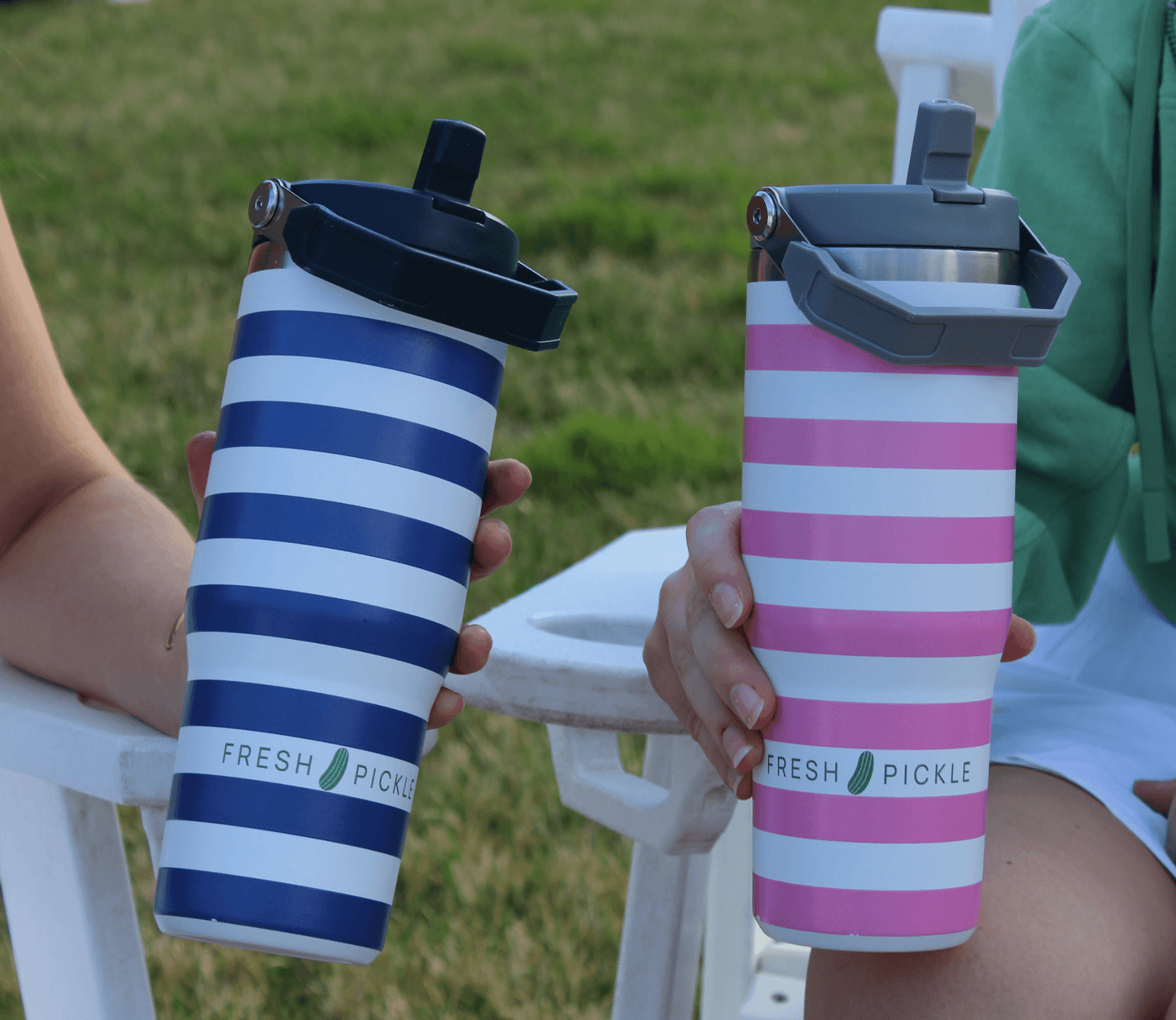 Two people sit on white outdoor chairs, each holding a Fresh Pickle Tumbler by Fresh Pickle. The 30 oz vacuum-sealed bottles, in blue/white or pink/white with a pickle graphic and flip-up straw, stand out against the grassy lawn backdrop.