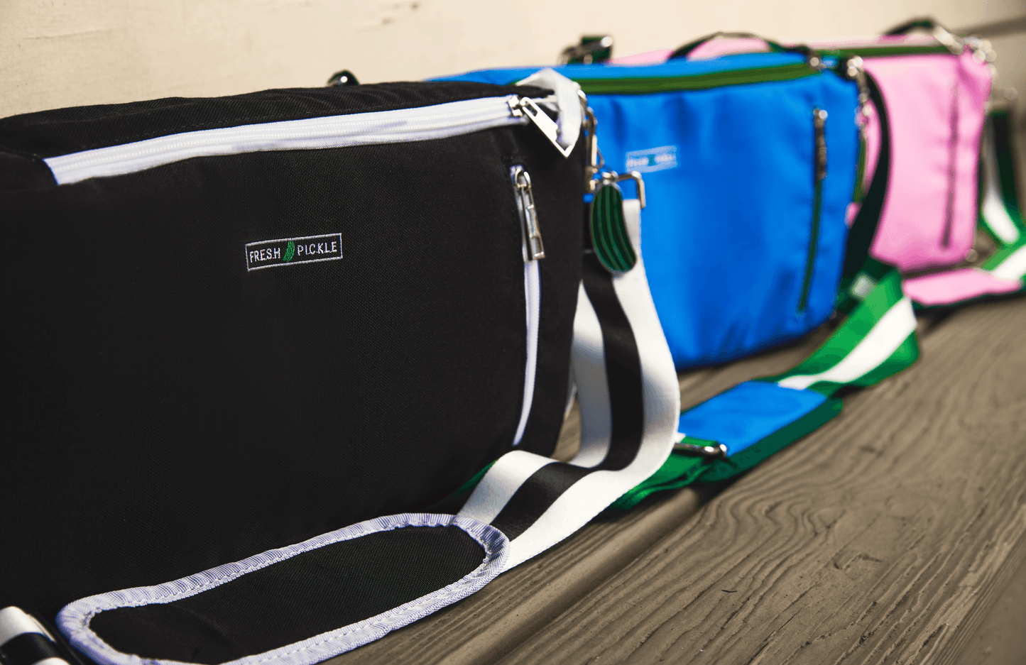 Three vibrant Insulated Cooler Bags by Fresh Pickle—in black, blue, and pink—are displayed with striped straps on wood. The front bag’s zip pocket and “Fresh Pickle” label add sporty style, perfect for carrying your court essentials.