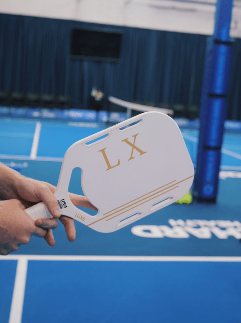 A person holds the lightweight, carbon fiber LUXE Pickleball LX paddle with gold “LX” letters on a USA Pickleball approved court featuring white lines, a net, blue post, curtains, and blurred signage in the background.