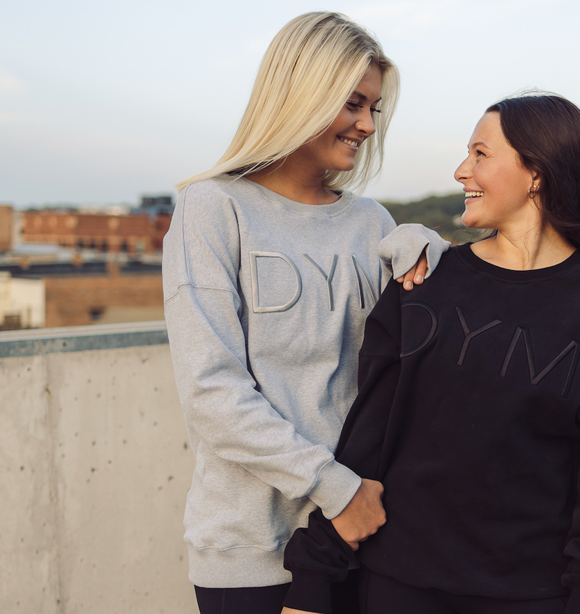 Two women smile at each other outdoors, wearing matching DYM Athletics Cozy Up Crew Neck Sweatshirts. With a relaxed fit and ultra-soft fabric, they look happy and comfortable against a backdrop of blurred buildings.